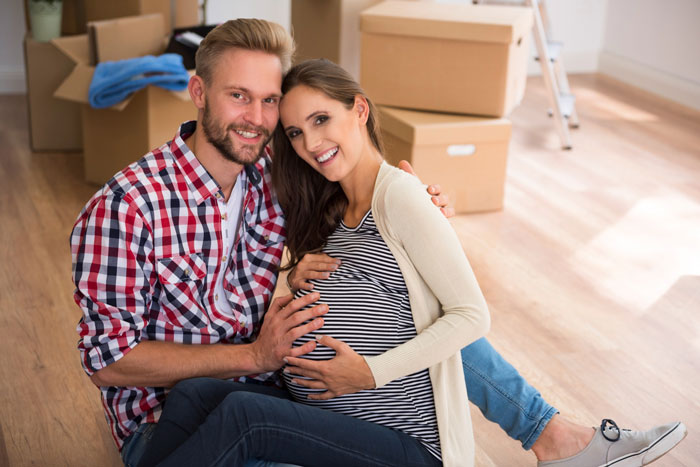 New parents, a pregnant woman and a man, smiling on the floor amidst moving boxes, disturbed by a rude neighbor. New parents, a pregnant woman and a man, smiling on the floor amidst moving boxes, disturbed by a rude neighbor.