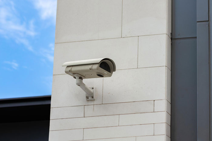 A security camera on a light-colored building with a blue sky, relevant to a guy fearing for his life due to eerie neighbor behavior.