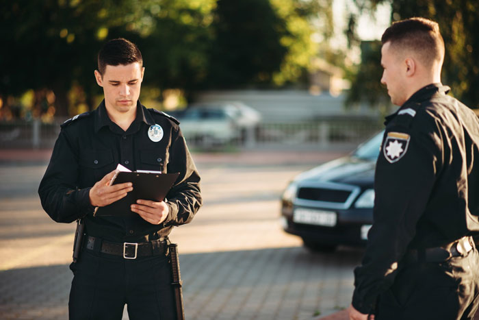 Two police officers in uniform stand outside, one looking down at a clipboard, while a homeowner fears for his life due to neighbor's eerie behavior.