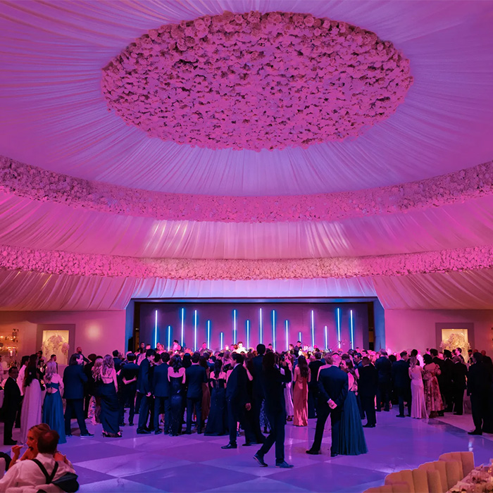 An extravagant wedding scene with guests on a dance floor illuminated by pink and purple lights. An extravagant wedding scene with guests on a dance floor illuminated by pink and purple lights.