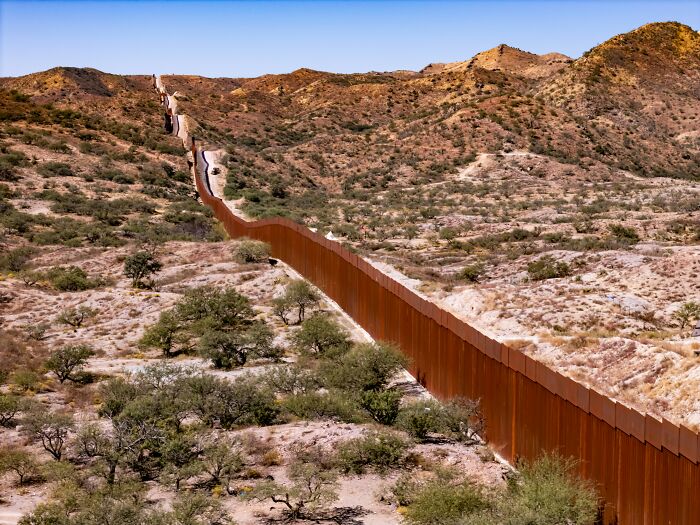 A long border wall stretches across arid, hilly terrain, surprising newcomers to America.