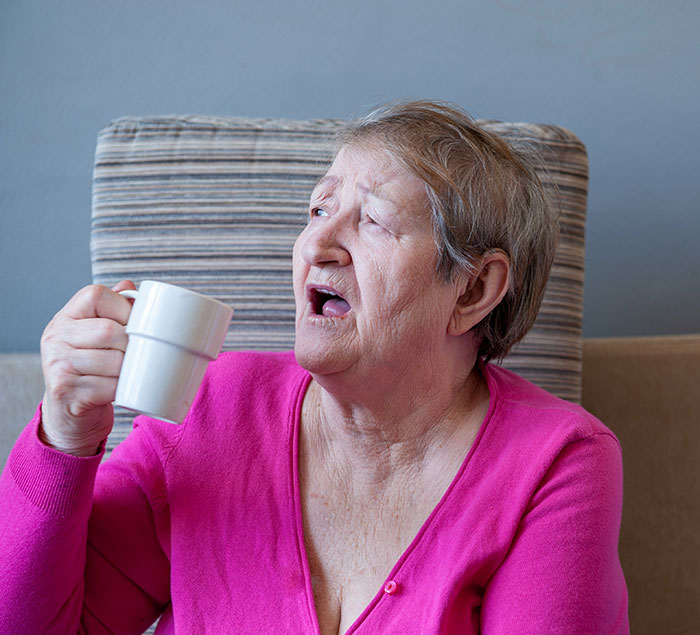Elderly woman with a displeased expression holds a white coffee mug, reflecting the MIL's refusal.