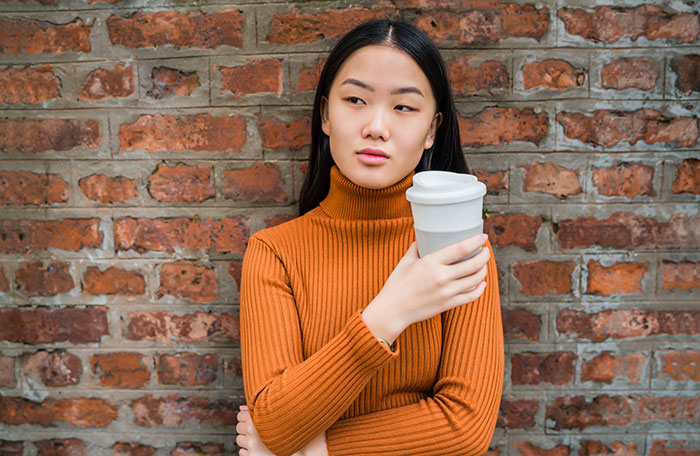 A woman holding a reusable coffee cup, looking thoughtful against a brick wall, relating to coffee order.