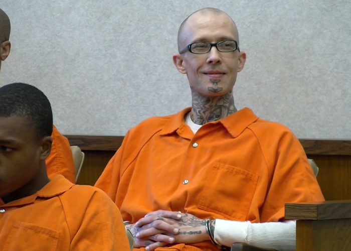 A man with tattoos on his neck and hands, wearing an orange jumpsuit, smiling while looking to the right. A man with tattoos on his neck and hands, wearing an orange jumpsuit, smiling while looking to the right.