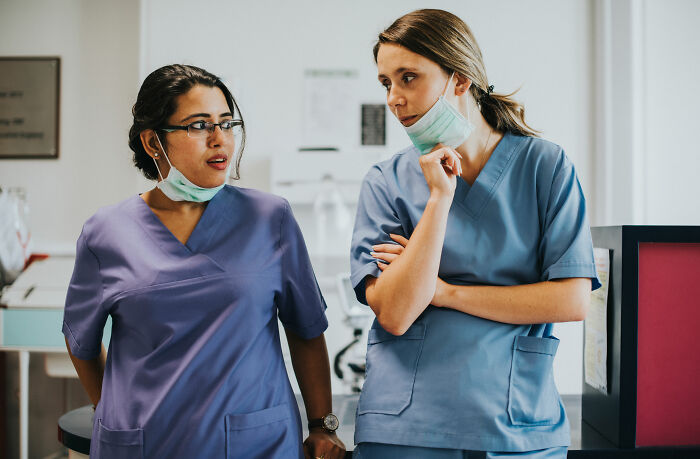 Two coworkers in scrubs and masks, discussing personal details.