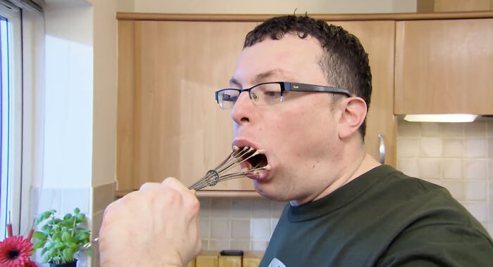 A man with glasses putting a whisk in his mouth, an example of dry British humor.