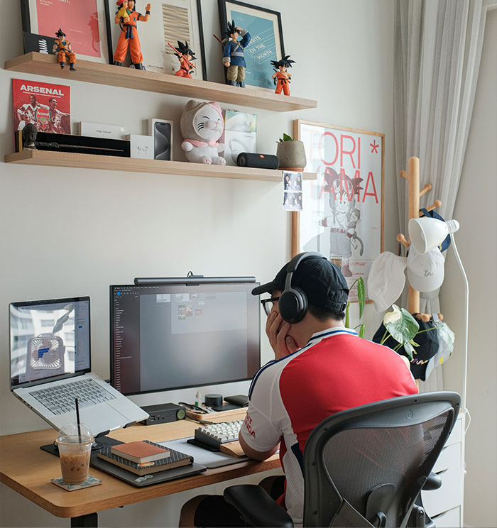 A young man with headphones, working on a computer in a tidy room with shelves, reflecting on the serious talk.