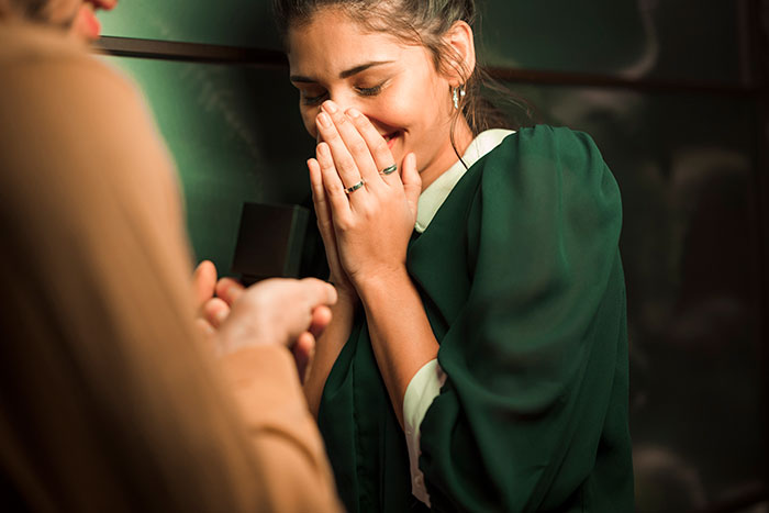 A GF looks surprised and happy, covering her mouth, as her BF proposes to her, showcasing female friends.