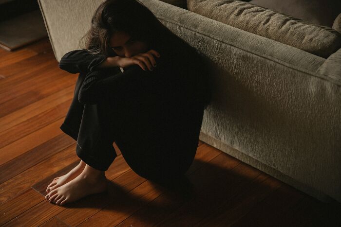 A person sits curled up on a wooden floor next to a couch, their face obscured, evoking the scariest things people witness.