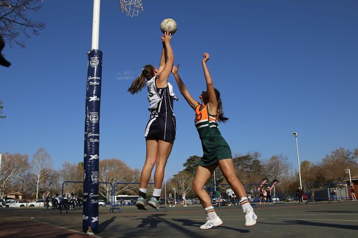 Two female athletes in mid-air, one shooting for the netball hoop while the other defends, showing competitive gender roles.