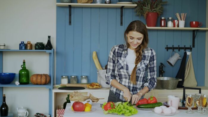 A woman in a plaid shirt is smiling while chopping vegetables in a kitchen. This image relates to toxic femininity.