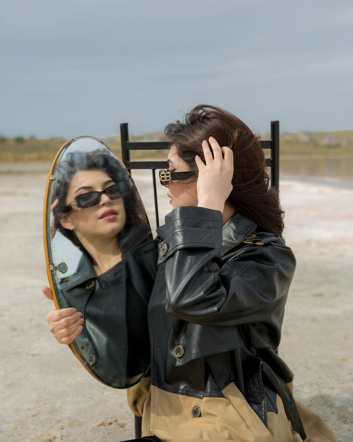 A woman in a leather jacket and sunglasses holding a mirror, examining her reflection, exploring themes of femininity.