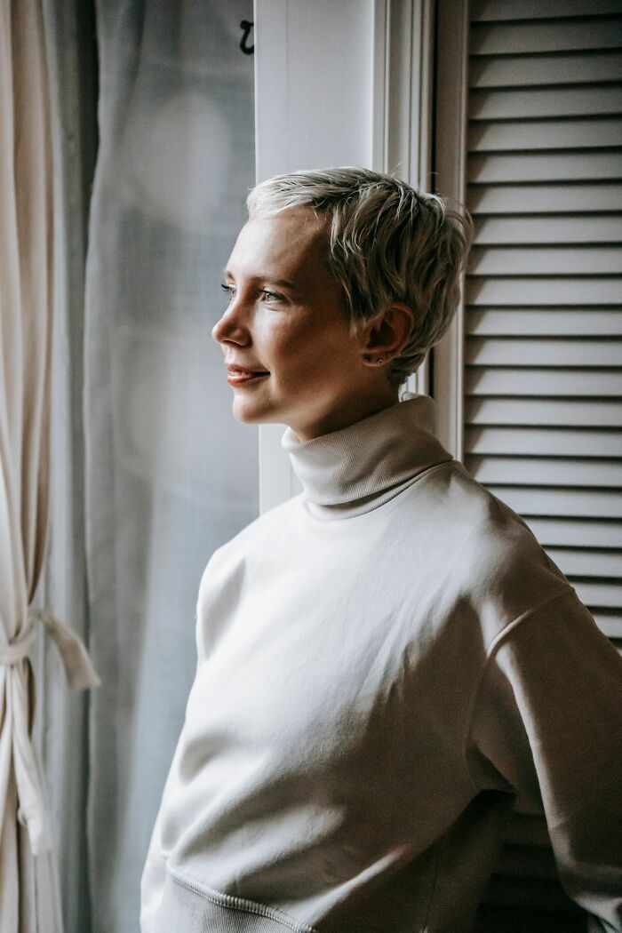 A woman with short blonde hair in a white turtleneck looking out a window, reflecting on gender roles and toxic femininity.