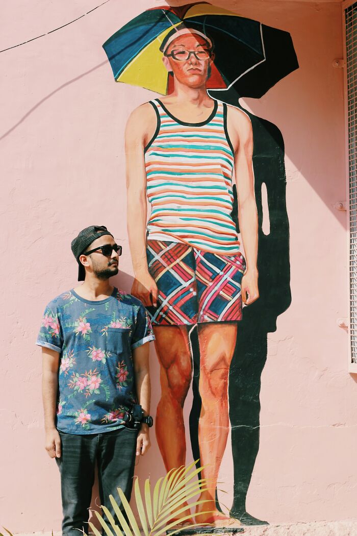 A man in a floral shirt stands next to a mural of a larger man, illustrating gender roles.