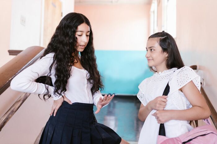 Two girls in school uniforms are talking in a hallway, one with a backpack. This image relates to toxic femininity.