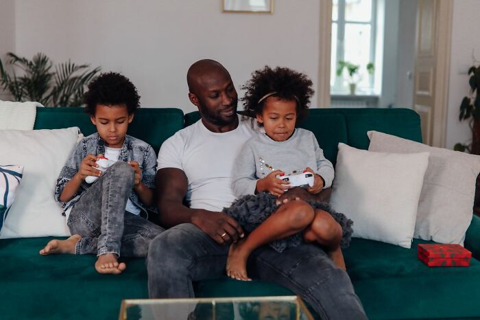 A father sits on a couch with his two children, who are playing video games. This image relates to toxic femininity.