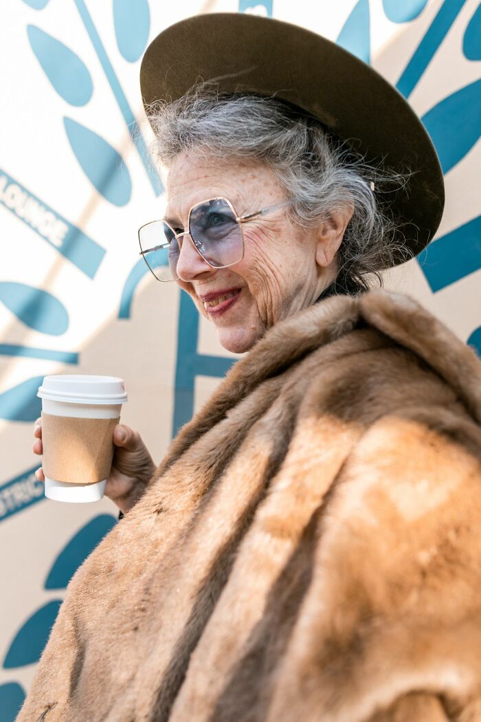 An older woman in a fur coat, hat, and sunglasses holding a coffee, smiling, embodying a confident expression of femininity.