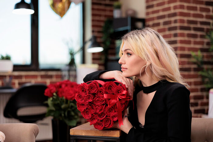 A woman with blonde hair gazes away from a heart-shaped box of roses, hinting at affairs coming to light brutally.