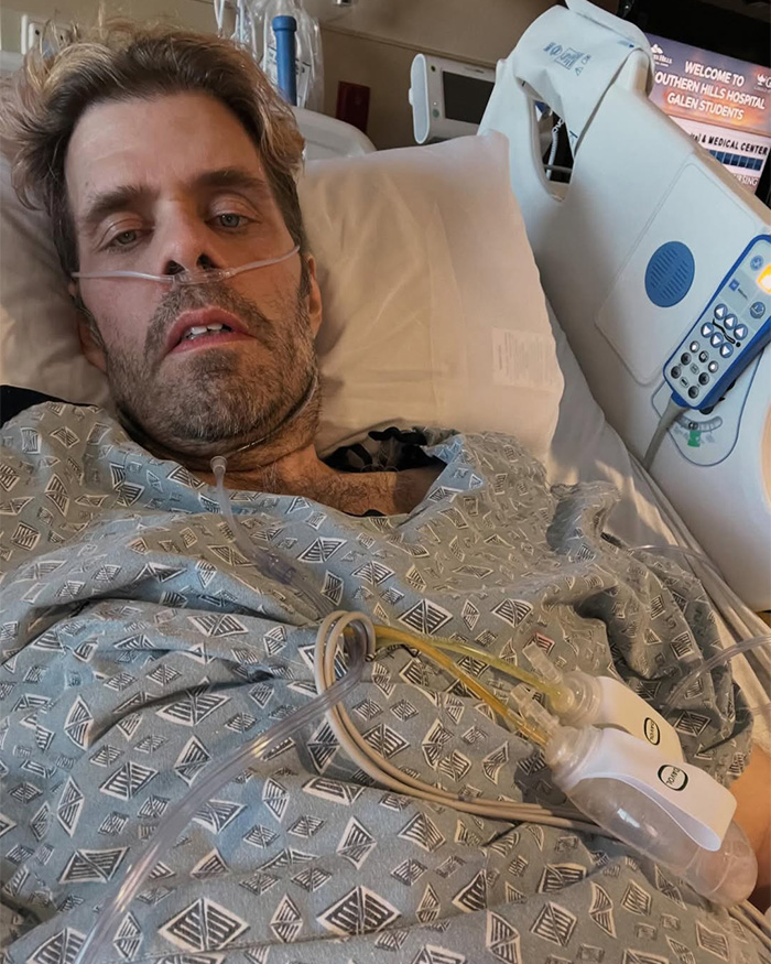 Perez Hilton in a hospital bed, connected to medical equipment, looking directly at the camera with a serious expression.