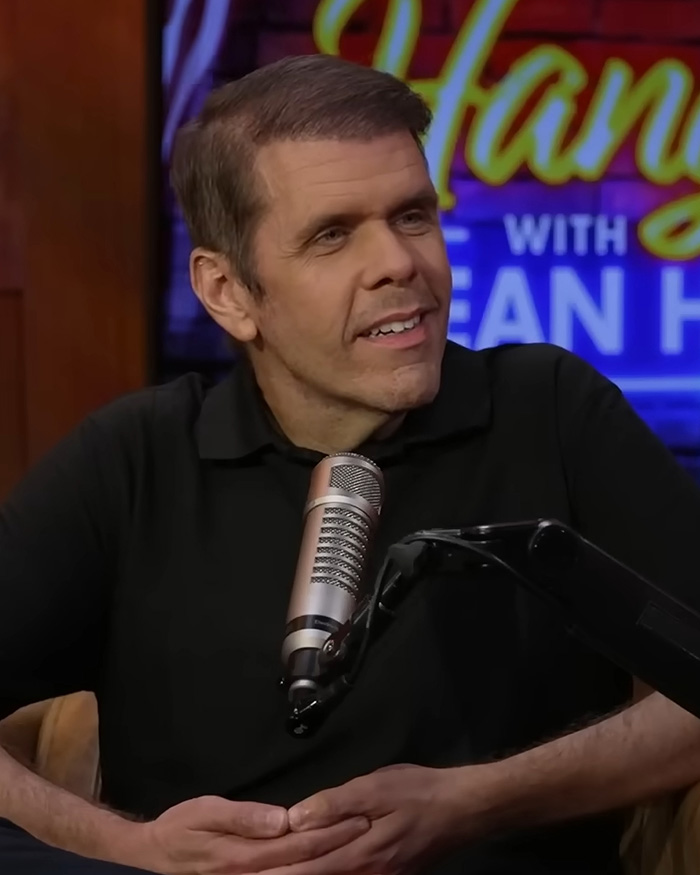 Perez Hilton, smiling, in a black polo shirt, with a microphone in front of him, for a podcast or interview.