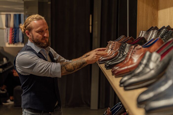 Man with tattoos adjusting a pair of brown leather shoes on a shelf, showcasing sick burns of style.