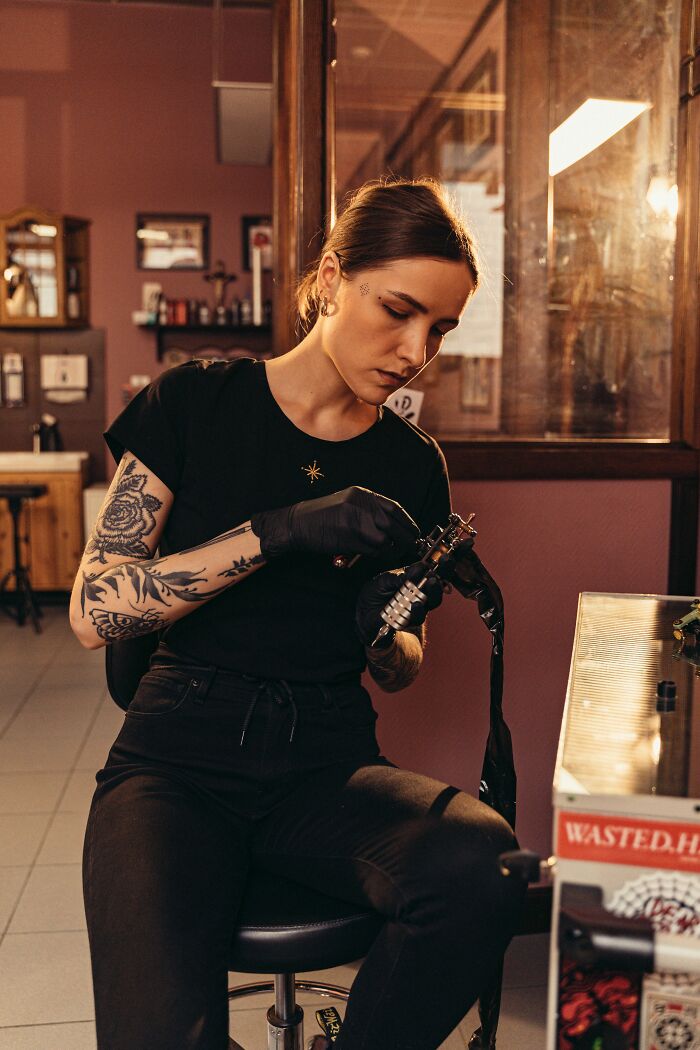 A female tattoo artist with tattoos on her arms, wearing gloves and preparing a tattoo machine, showing personal details of coworkers.