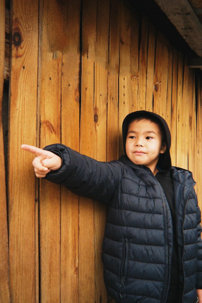 A young boy in a black puffer jacket pointing to the left, standing against a wooden wall, for creepy true stories.