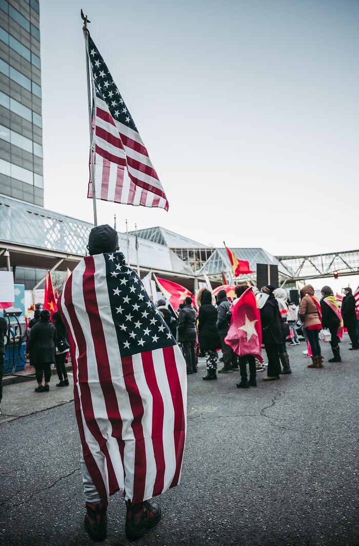 A person with an American flag draped over their back, standing in a crowd, symbolizing beliefs and propaganda.