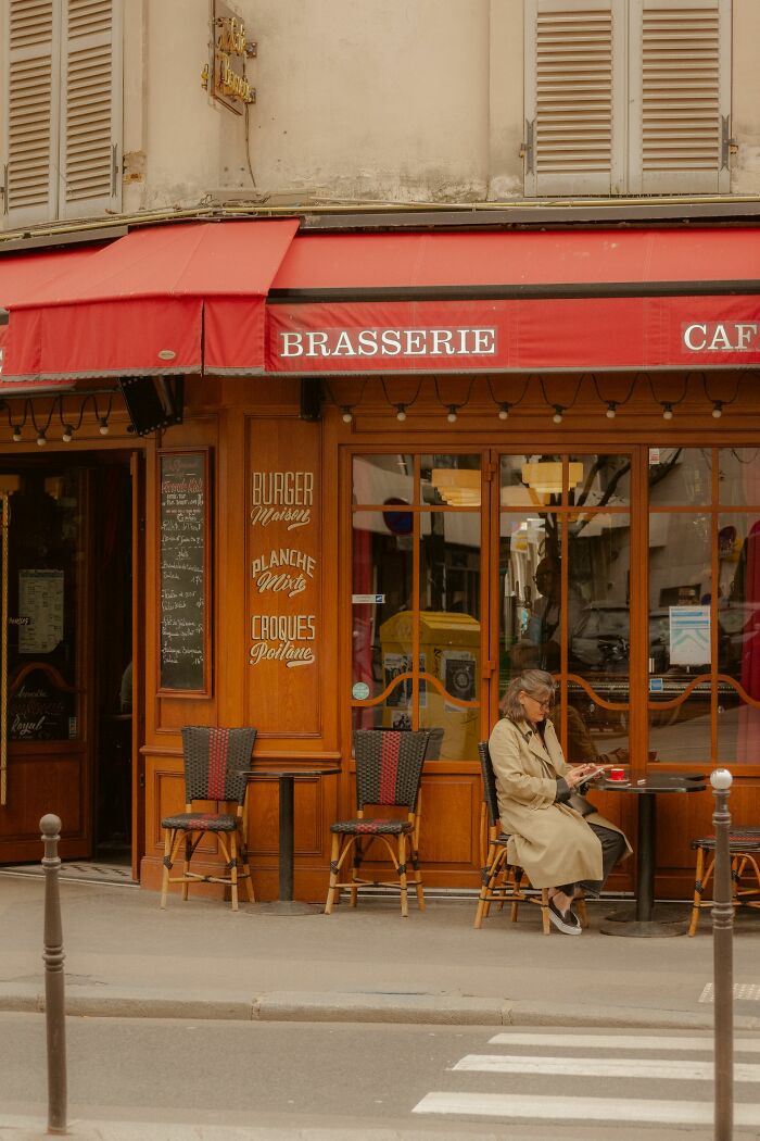 A woman sitting at an outdoor table of a French brasserie under a red awning. People believe today that propaganda influences beliefs.