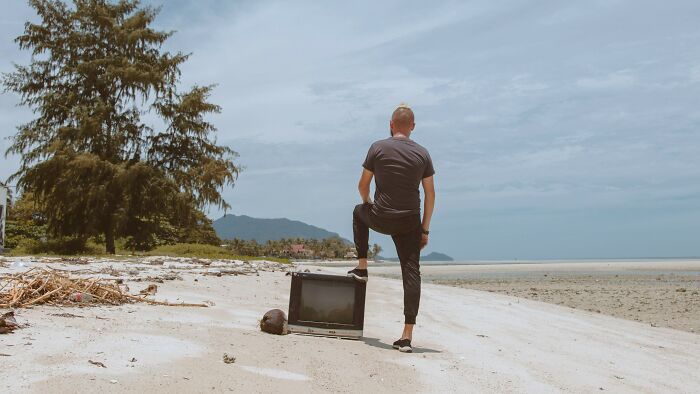 A man stands on a beach with one foot on an old TV, looking out at the ocean, representing reality TV questions.