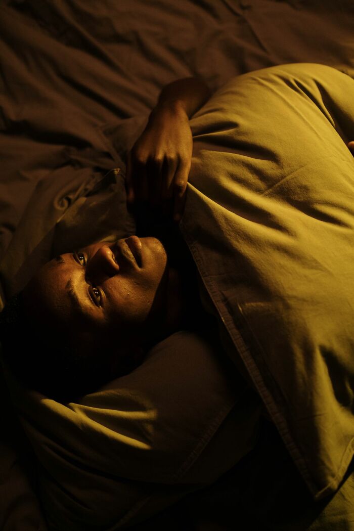 A man lies awake in bed, his face lit by a dim light, looking unsettled by creepy true stories.