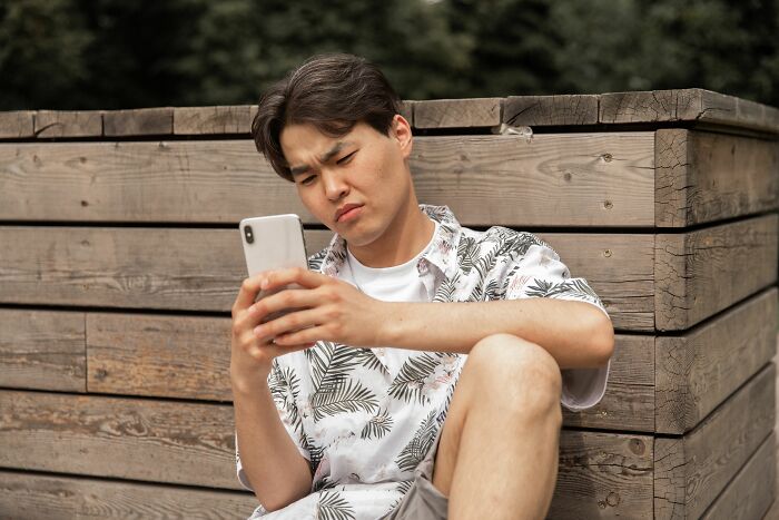 A man sitting outdoors, frowning at his phone, surrounded by wood planks.