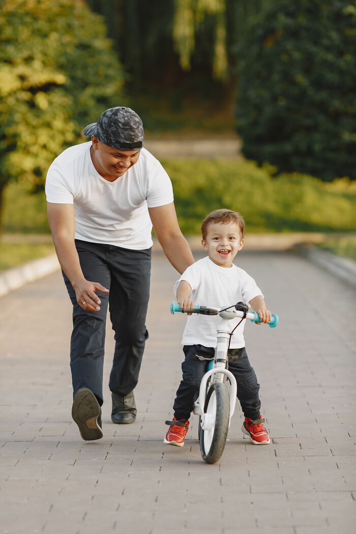A man helps a small child ride a balance bike on a paved path in a park, a common sight in America.