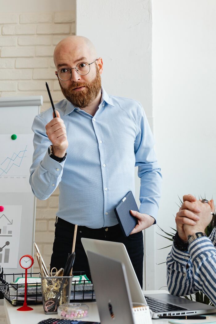 A man with a beard and glasses stands in an office, holding a pen, ready to deliver a sick burn.