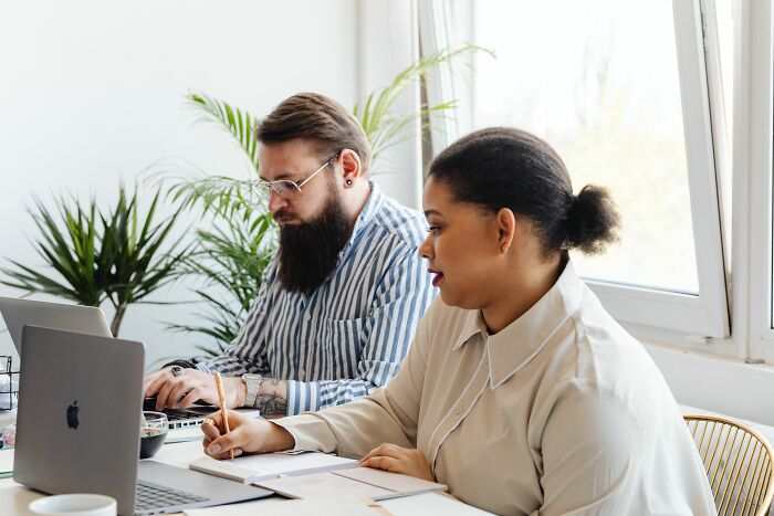 Two coworkers, a man with a beard and a woman, working on laptops and taking notes, focusing on coworker interactions.