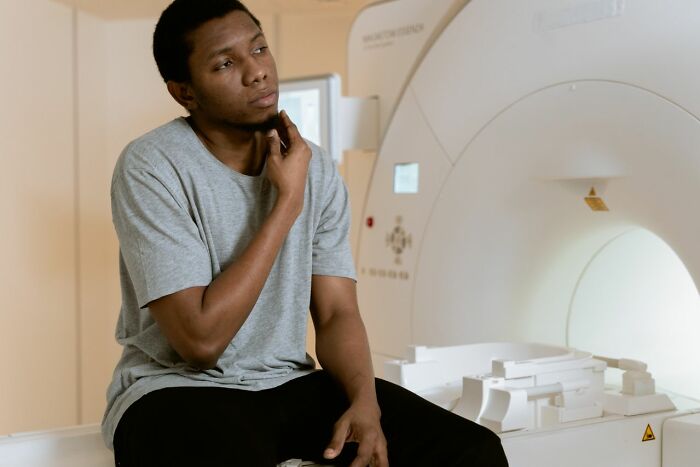 A young man in a medical setting, possibly a patient or medical field worker, sits contemplatively beside an MRI machine.