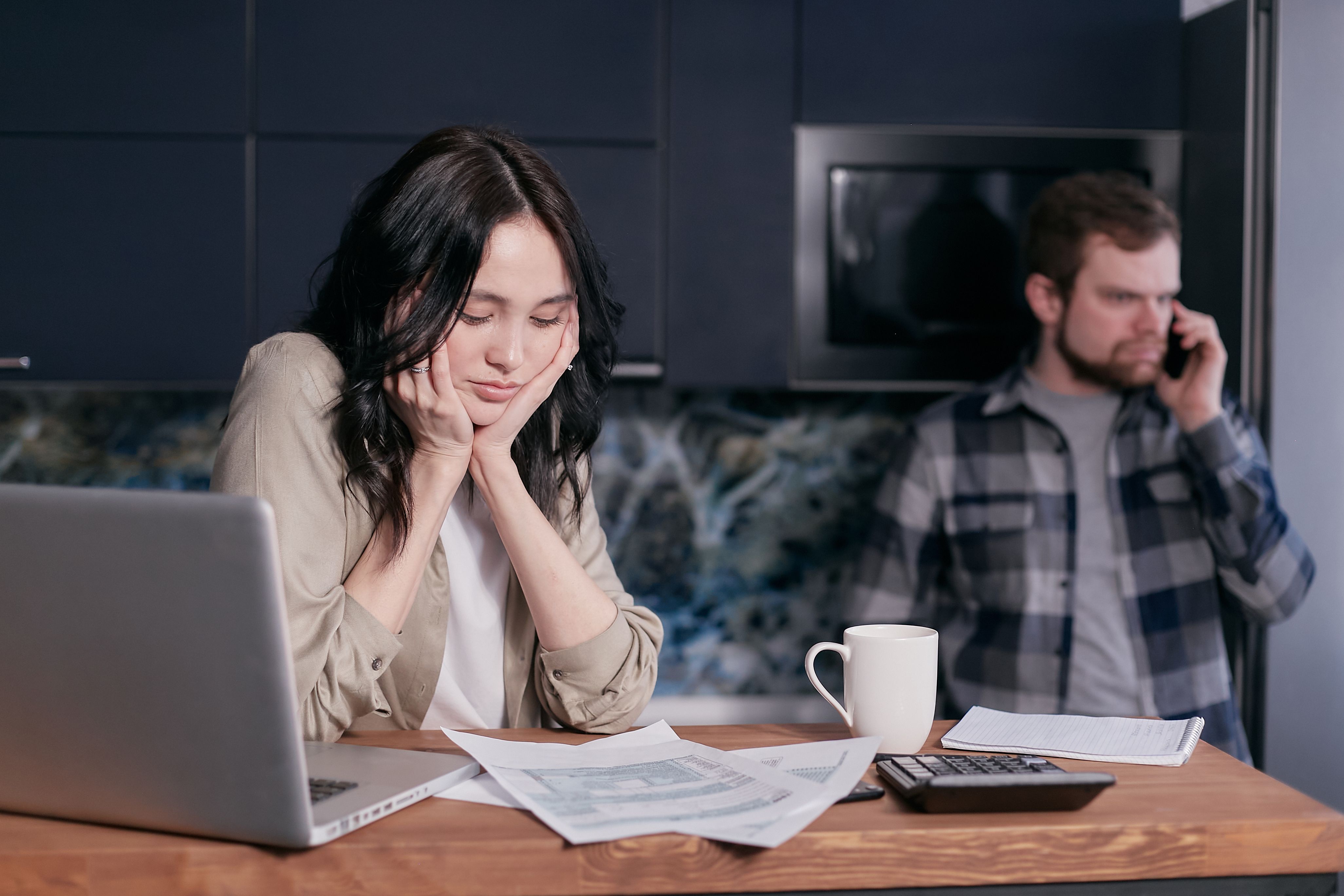 A frustrated woman at a laptop with papers, while a man talks on the phone in the background, showing eviction stress. A frustrated woman at a laptop with papers, while a man talks on the phone in the background, showing eviction stress.