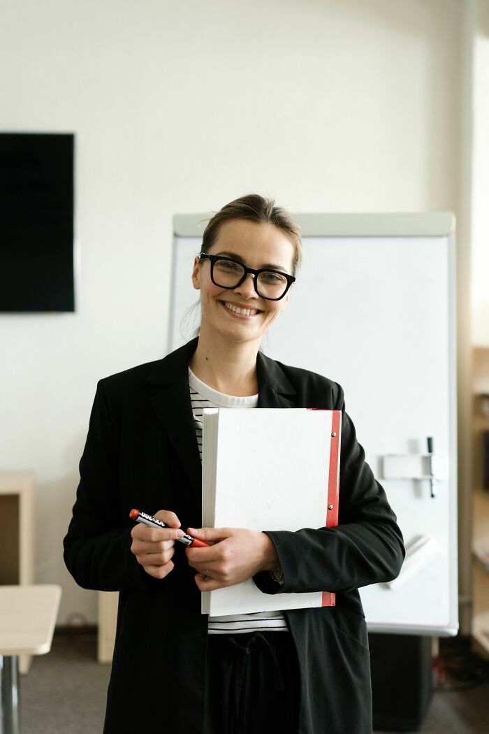 A smiling woman in glasses and a blazer holding a notebook and marker, symbolizing incredible accomplishments.