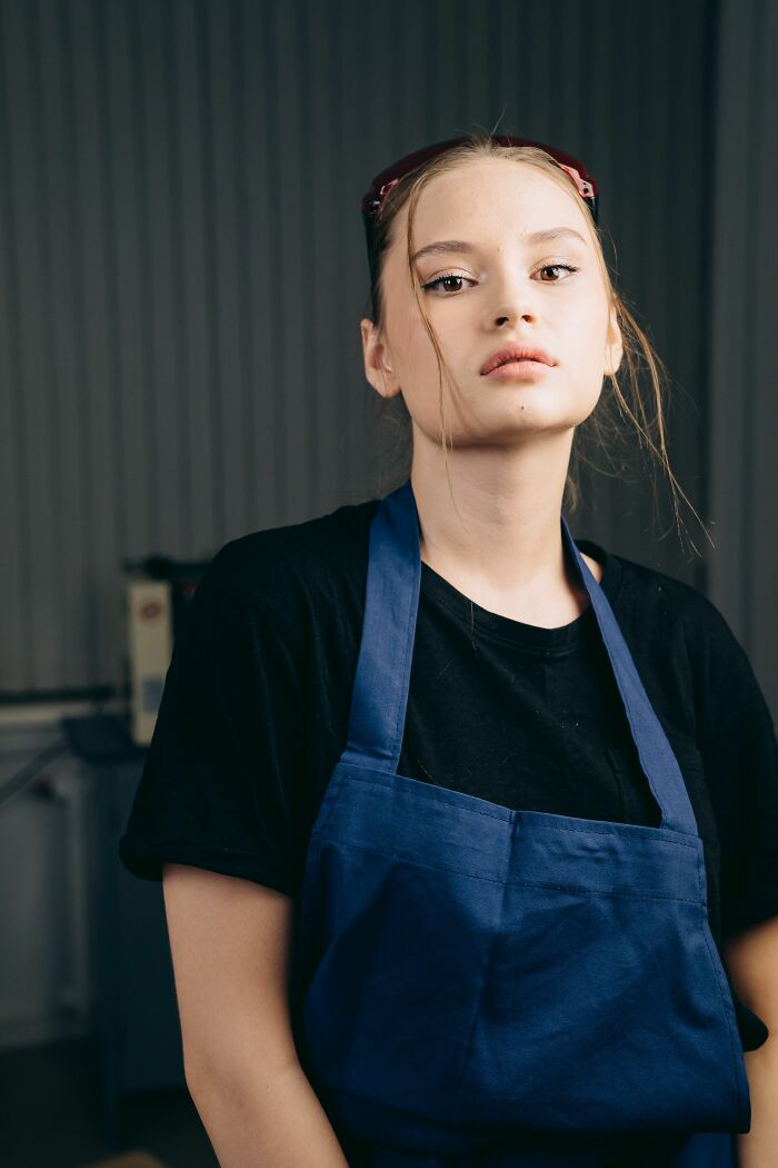 A young woman in a black shirt and blue apron, looking intensely.