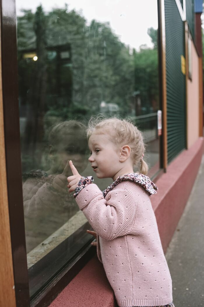 Little girl with blonde pigtails pointing at her reflection in glass, looking scared by the true story.