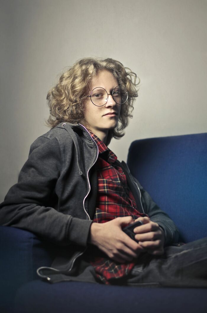 A young person with curly hair and glasses sits on a blue couch, delivering a sick burn.