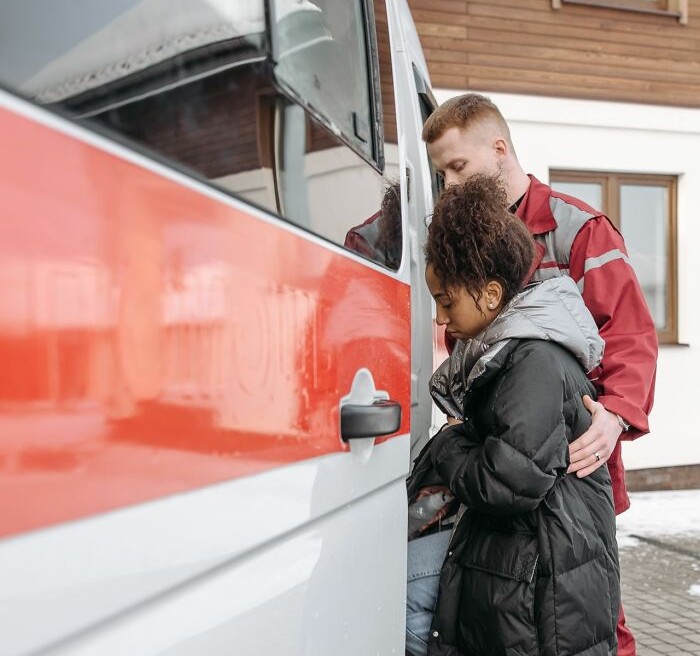 A medical field worker in a red uniform assists a distressed woman into an ambulance, highlighting patient care.