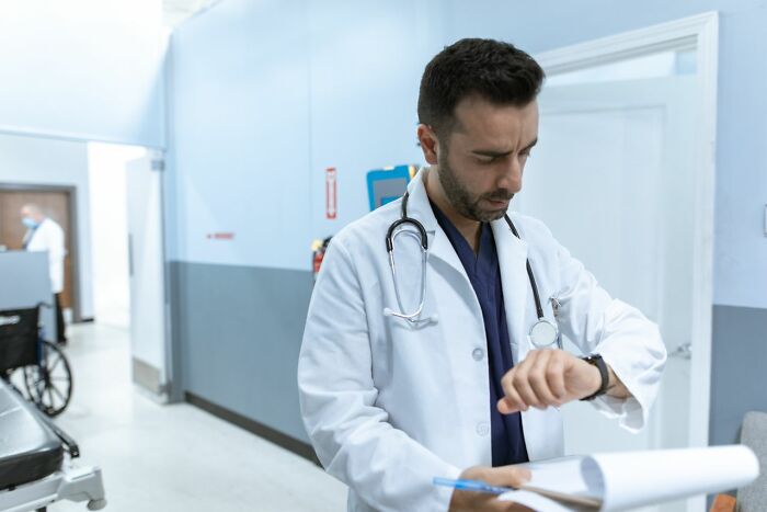 A medical field worker, dressed in a lab coat, checks his watch and holds a clipboard in a hospital hallway.