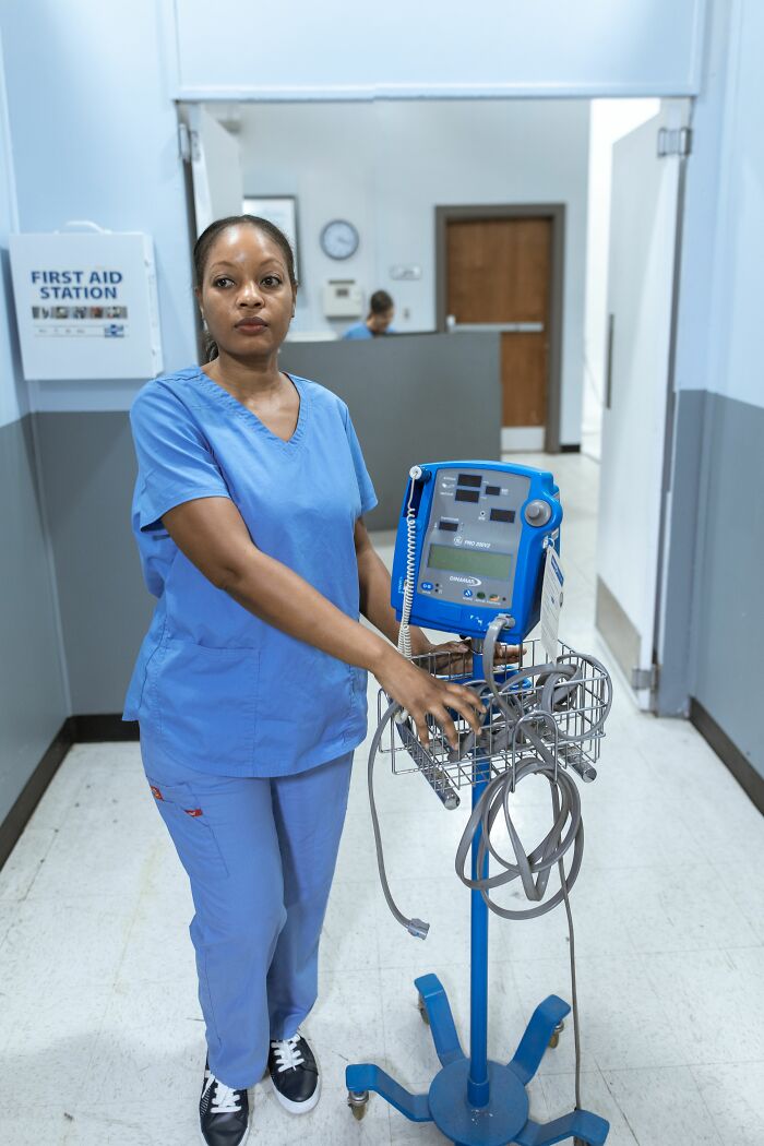 A nurse standing in a hospital hallway, pushing medical equipment, possibly affected by coworkers personal details and PTSD mode.