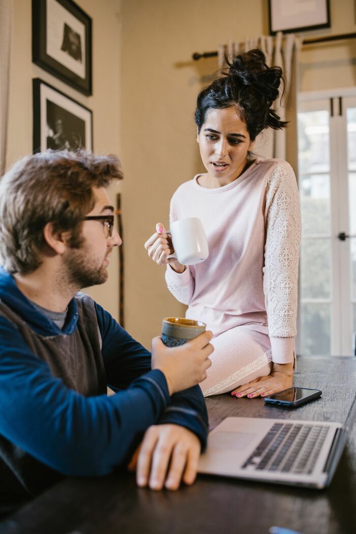 A woman sitting on a table with a mug looks distressed while talking to her boyfriend who is on a laptop.