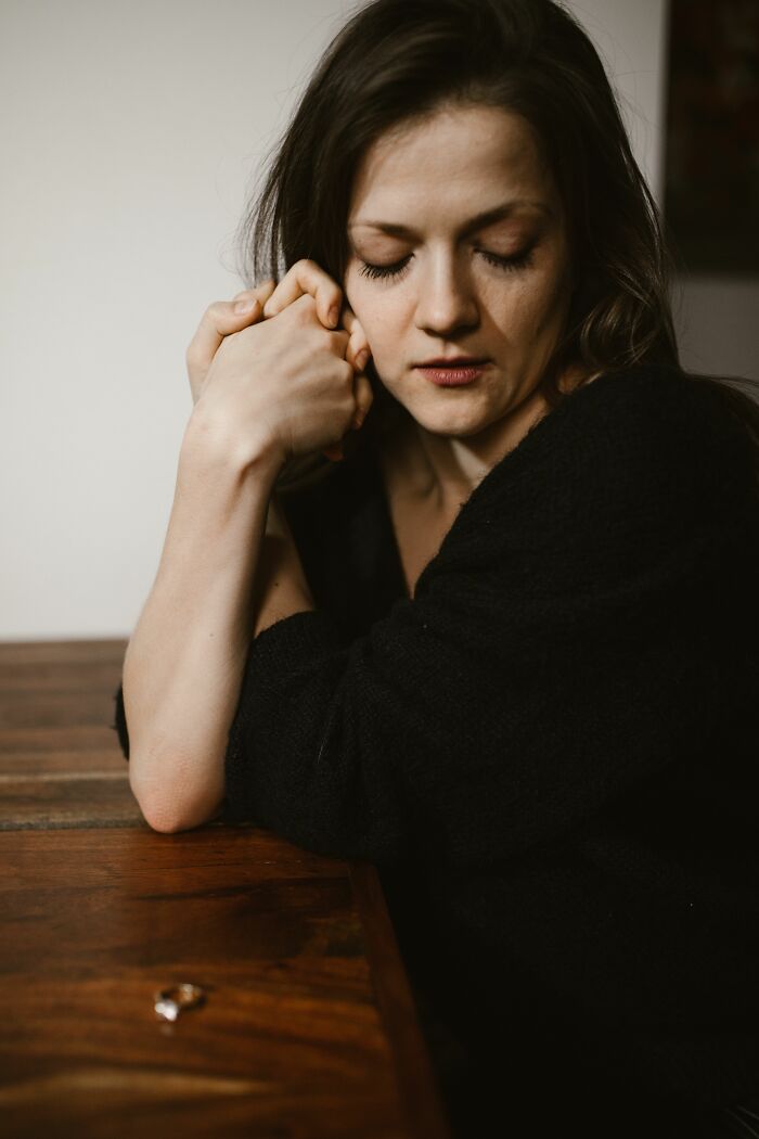 A woman with closed eyes and clasped hands, next to a wedding ring, realizing she is with the very wrong person.