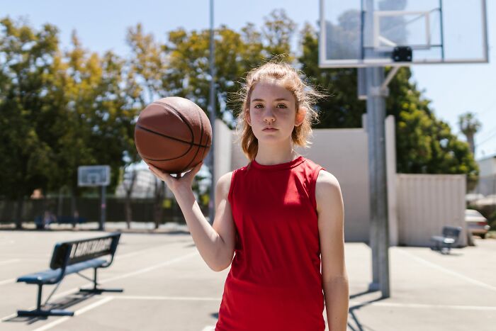 A young woman in a red shirt holds a basketball on a court, embodying the confidence of someone delivering sick burns.