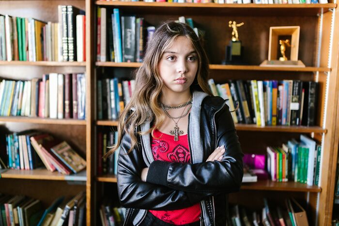 Young girl with long hair and a cross necklace standing in front of bookshelves, projecting sick burns of youthful defiance.