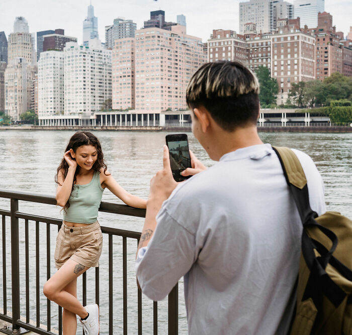 A man with a backpack photographs a young woman posing by a railing near a river with a city skyline in the background. A man with a backpack photographs a young woman posing by a railing near a river with a city skyline in the background.