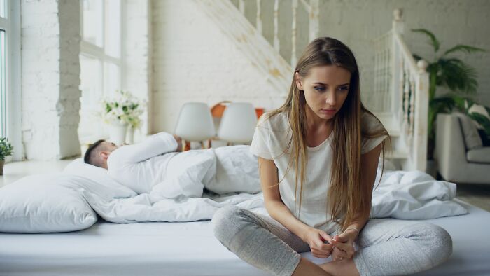 A woman sits on the bed, looking unhappy, while a man sleeps in the background, realizing she is with the very wrong person.